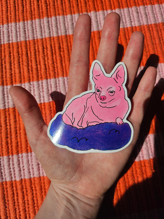 A die-cut sticker featuring a pink cartoon chihuahua sitting in a blue blanket, held in a person's hand over an orange and white striped knit blanket.