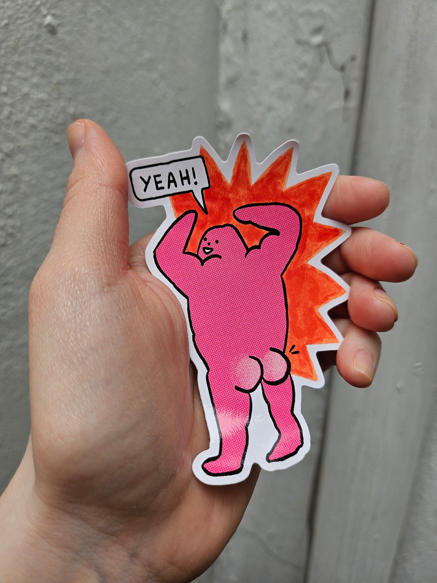 Hand holding a pink cartoon-style sticker with a character and 'YEAH!' text on a gray background