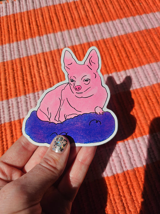 A hand with glittery nail polish holds a die-cut sticker of a pink chihuahua sitting in a blue blanket, set against a textured orange and white striped blanket.