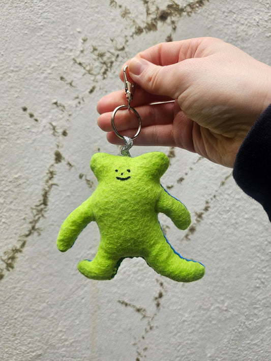 Green plush toy attached to a keychain held by a hand on a textured surface.