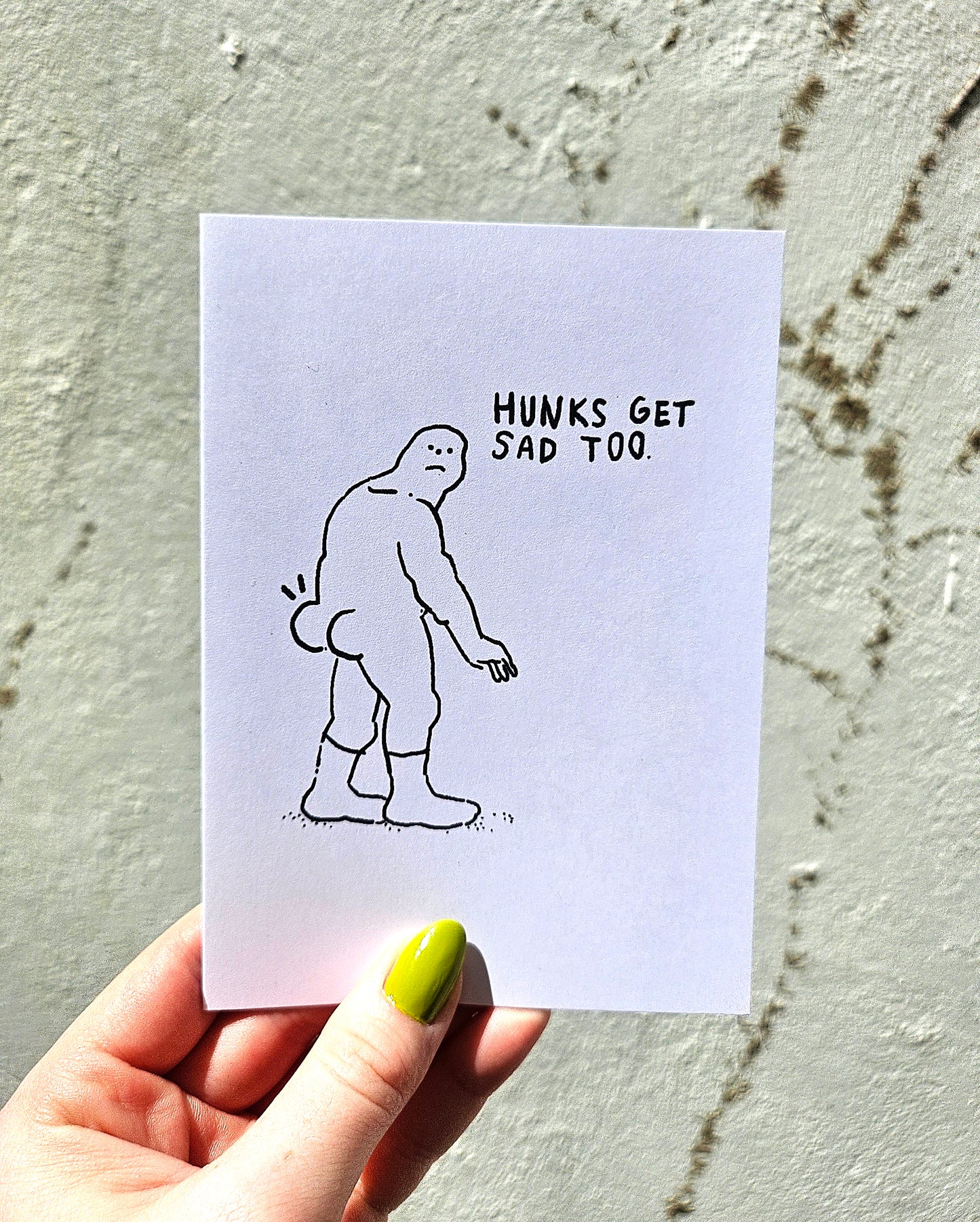 Hand holding a card with a cartoon of a sad figure and text 'Hunks get sad too' against a textured wall.