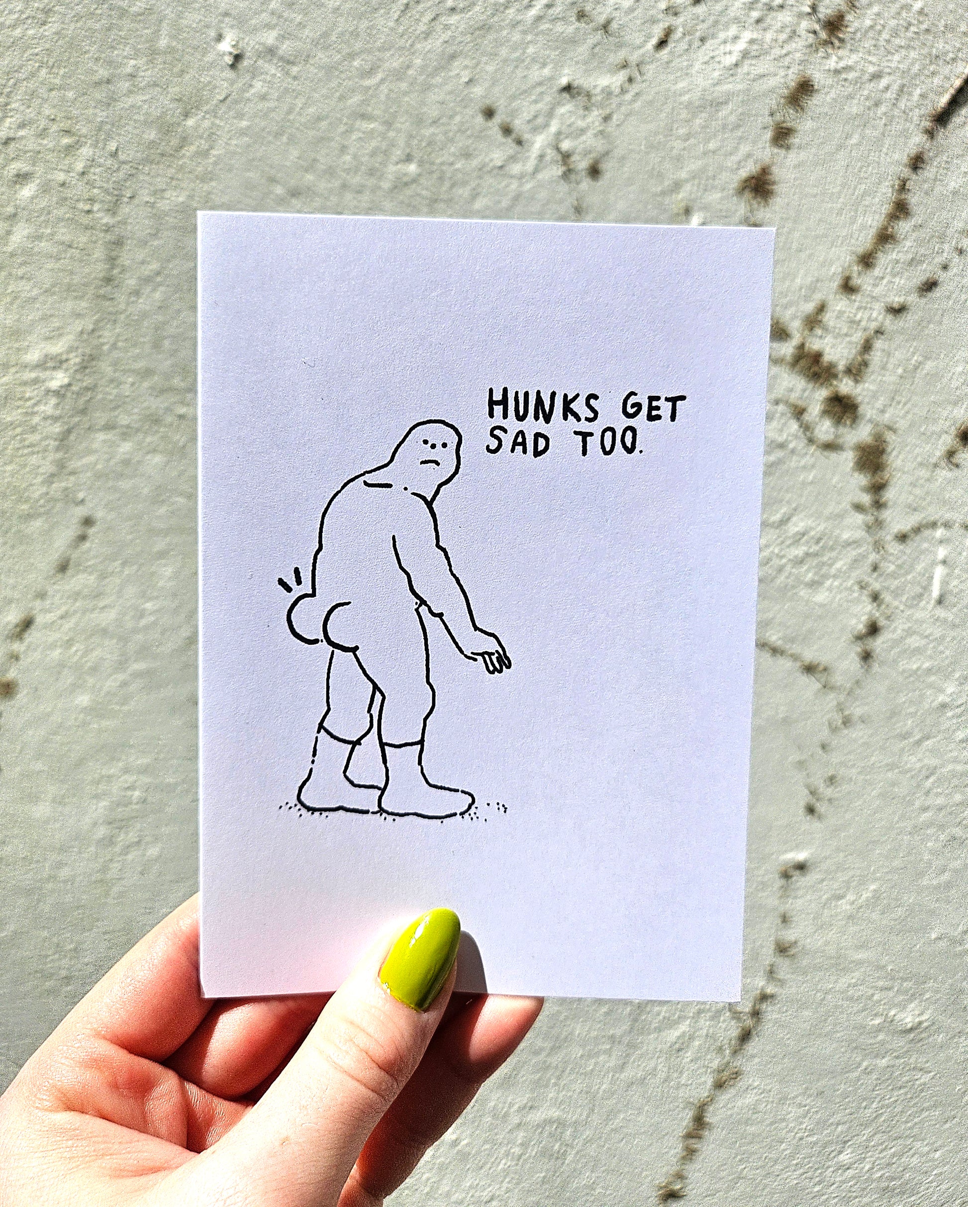 Hand holding a card with a cartoon of a sad figure and text 'Hunks get sad too' against a textured wall.