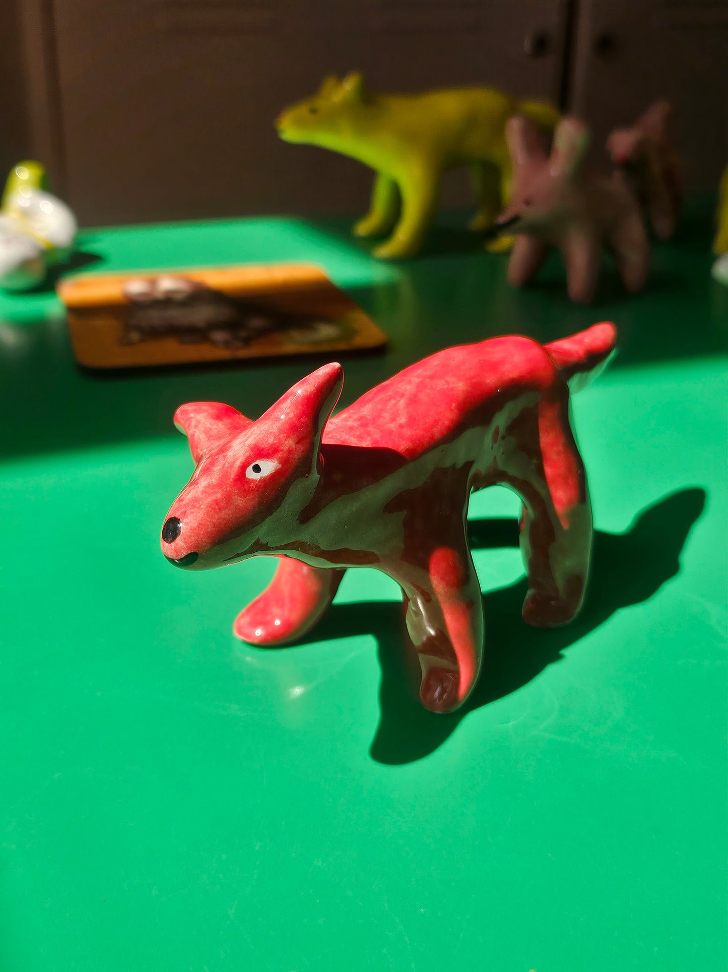A glossy red ceramic dog figurine standing on  a bright green surface, with other small figurines blurred in the background.