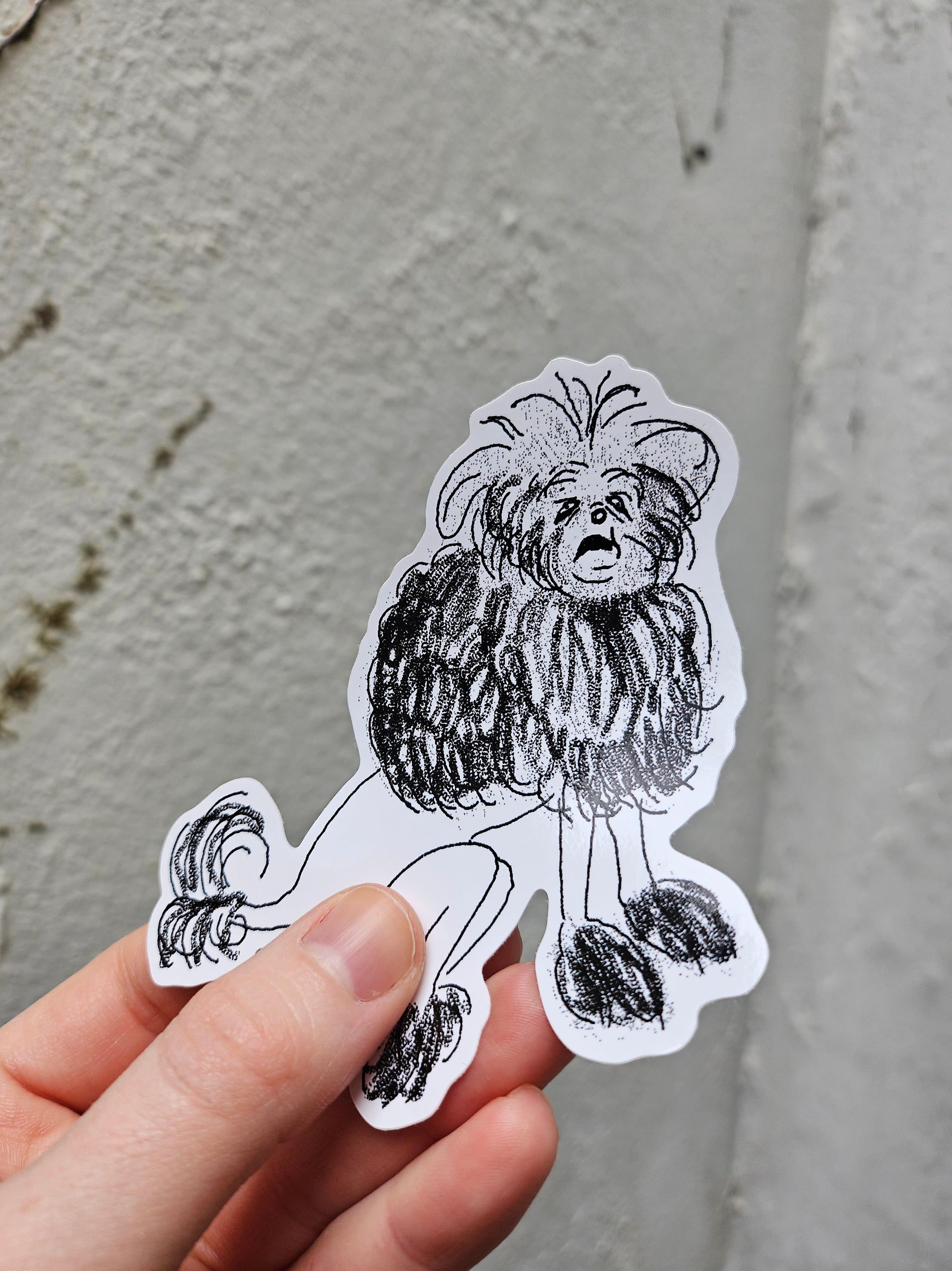 Hand holding a black and white illustrated sticker of an old, grumpy dog with long hair against a gray background