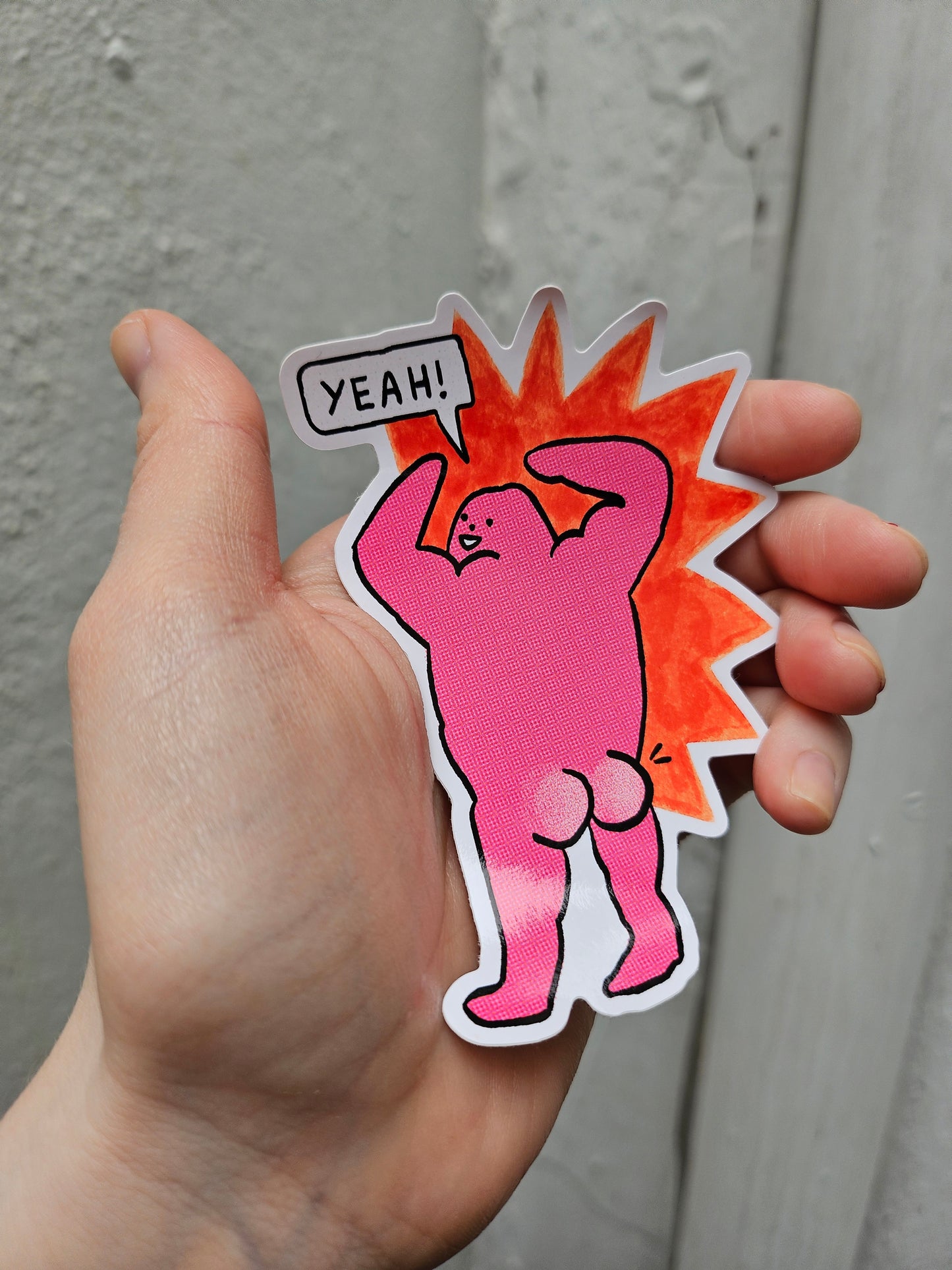 Hand holding a pink cartoon-style sticker with a sun and 'YEAH!' text on a neutral background