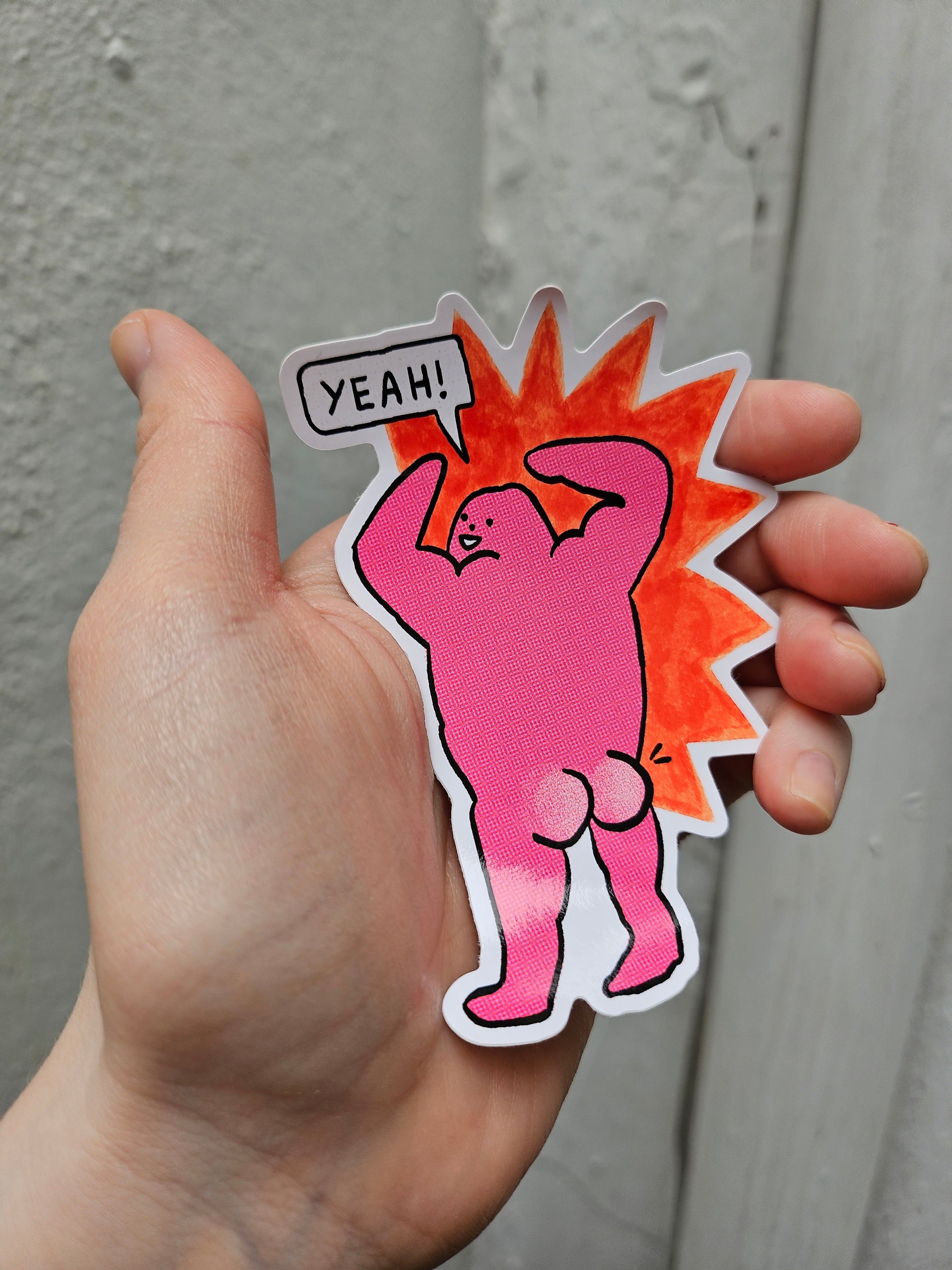 Hand holding a pink cartoon-style sticker with a sun and 'YEAH!' text on a neutral background