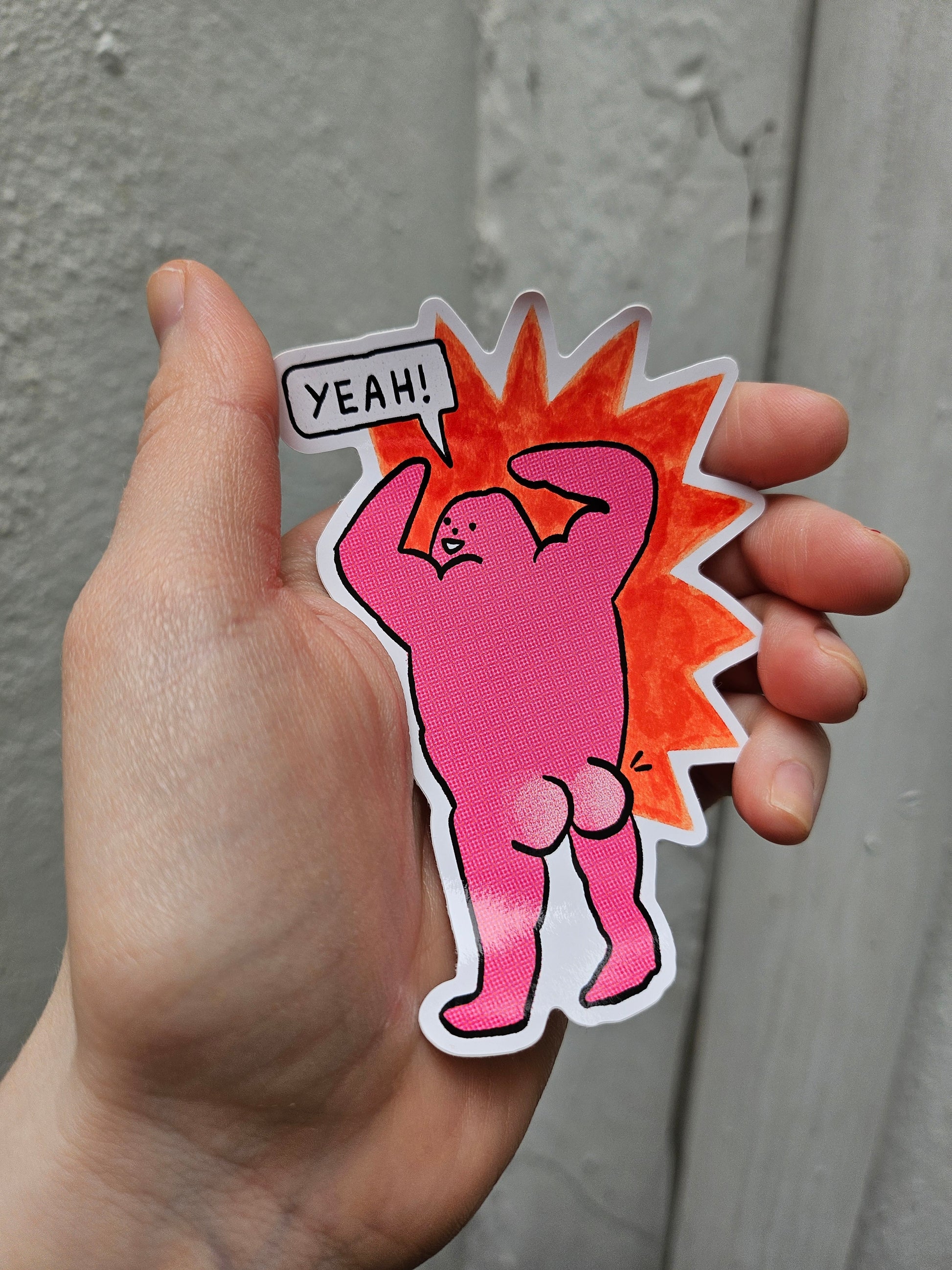 Hand holding a pink cartoon-style sticker with a character and 'YEAH!' text on a gray background