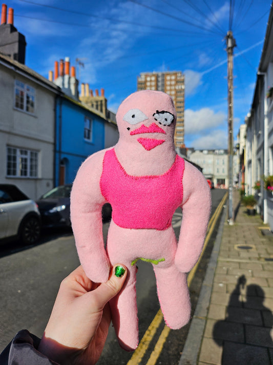 Pink plush toy held in front of a city street with buildings and blue sky.