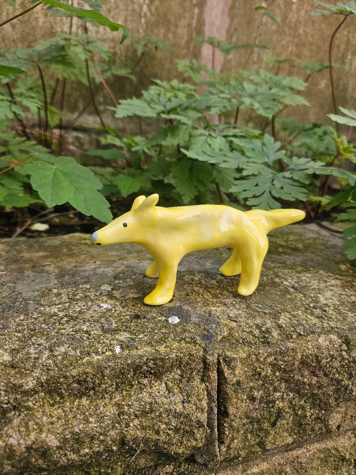 A small glossy yellow dog figurine mounted on a textured stone wall outdoors, with green leaves visible in the background.