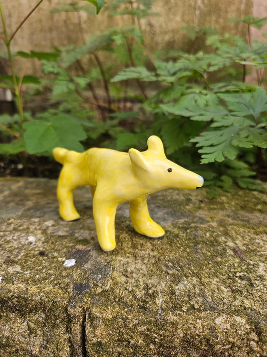 A small yellow ceramic dog figurine climbing on a textured stone wall outdoors with green plants in the background.