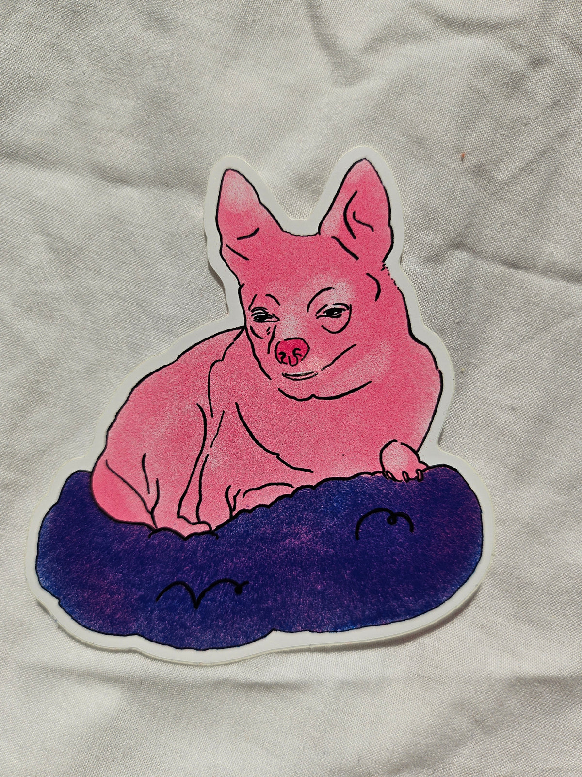 A die-cut sticker featuring a pink cartoon chihuahua lying on a dark purple cloud, placed on a wrinkled white fabric background.