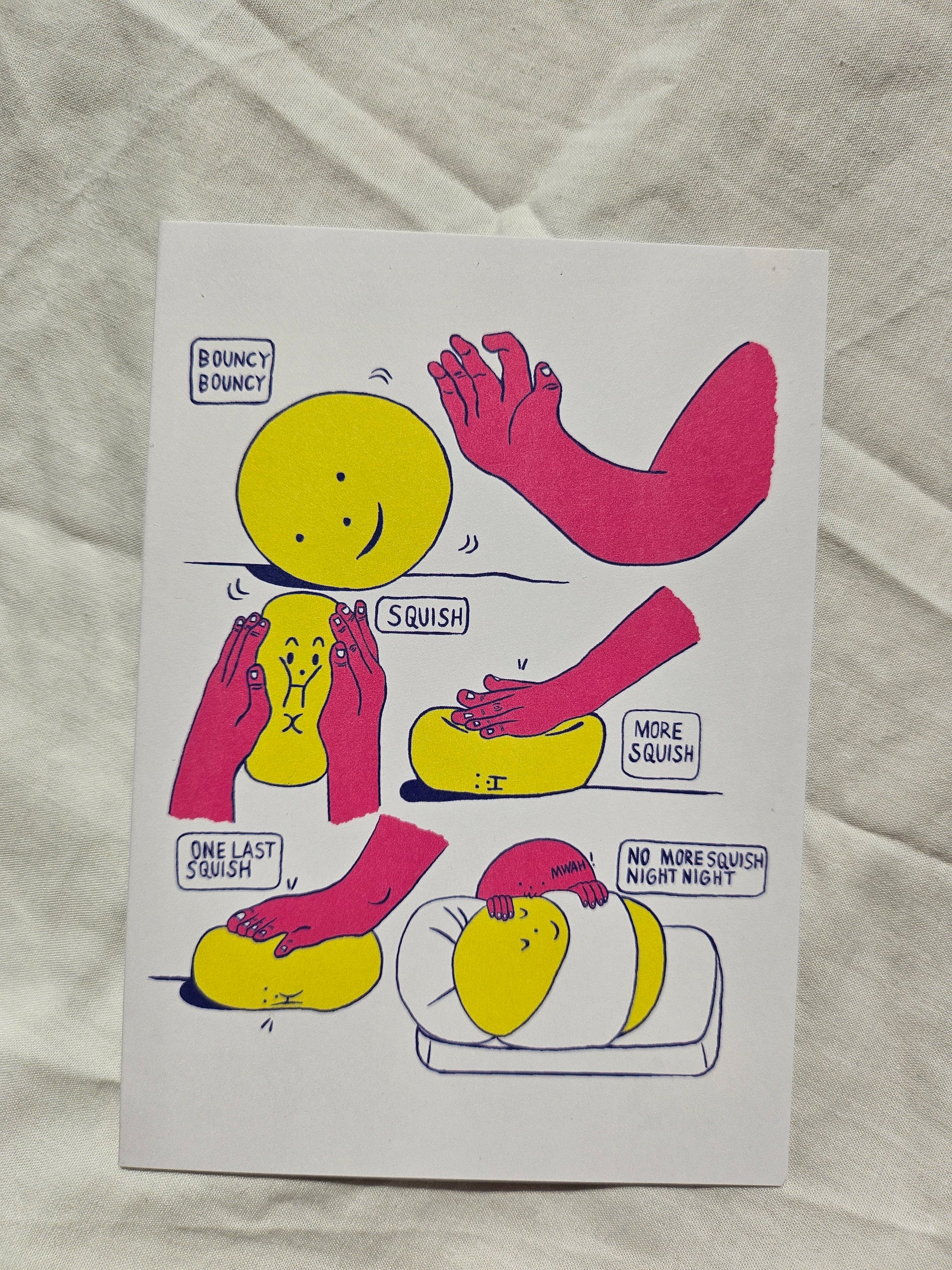 A white rectangular card featuring a comic-style illustration of yellow smiley faces and pink arms, placed on a wrinkled white fabric background.