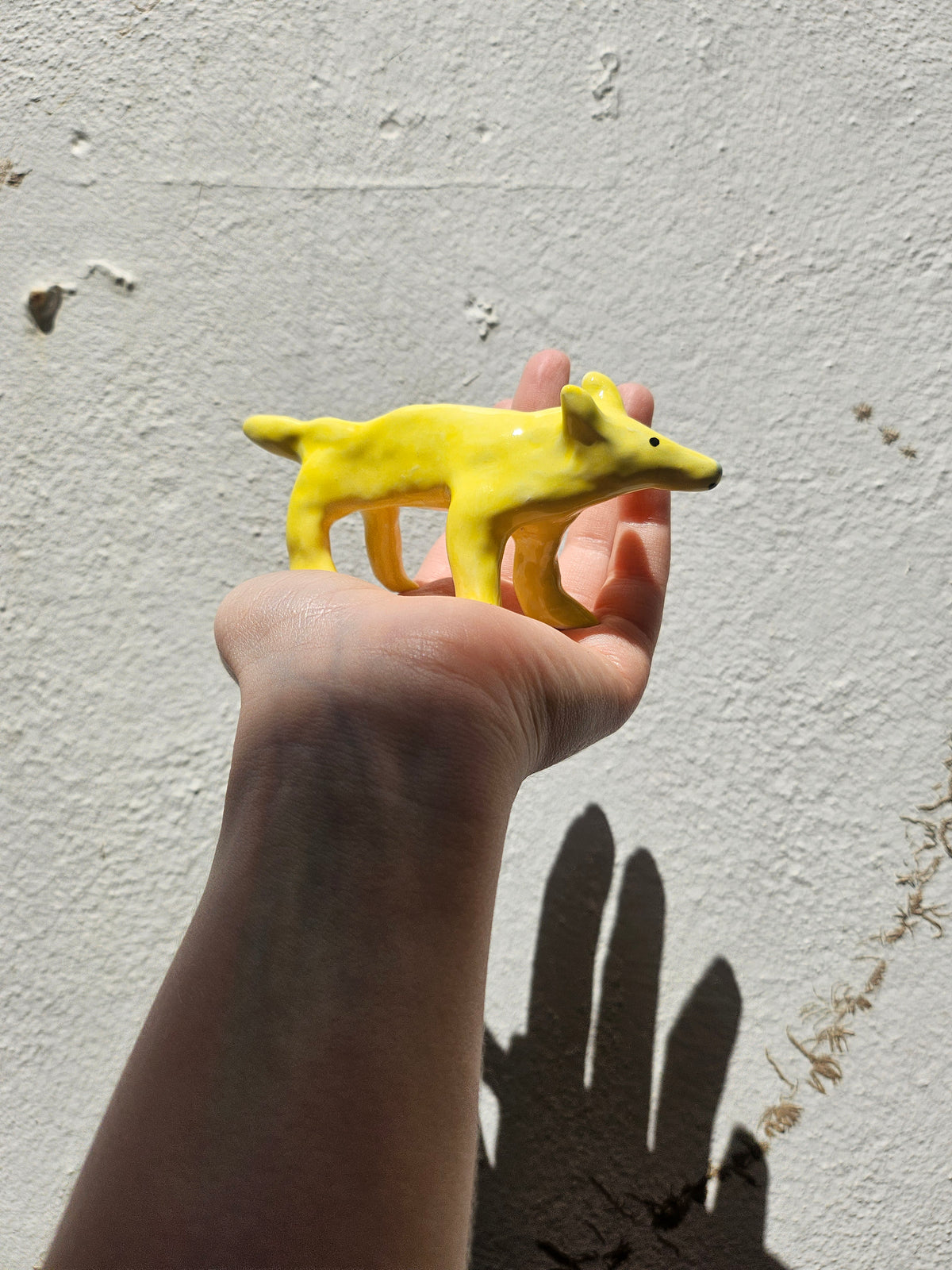 A hand holding a small yellow ceramic figurine of a dog against a textured off-white wall with a hand shadow.