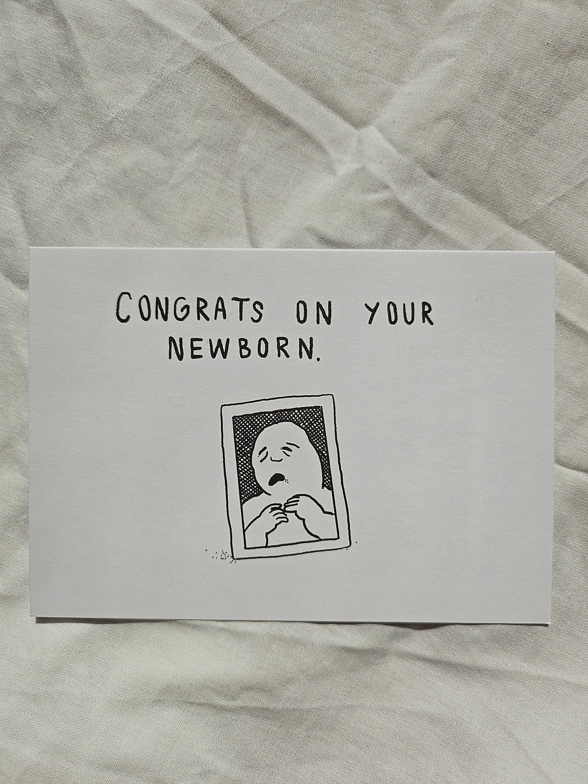 A white rectangle greeting card with black text reading 'CONGRATS ON YOUR NEWBORN.' and a simple black line drawing of a baby, placed on a wrinkled off-white fabric background.