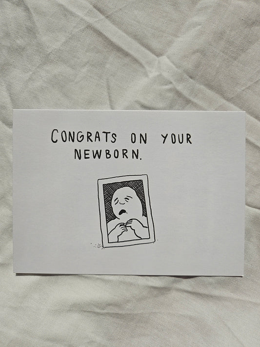 A white rectangle greeting card with black text reading 'CONGRATS ON YOUR NEWBORN.' and a simple black line drawing of a baby, placed on a wrinkled off-white fabric background.